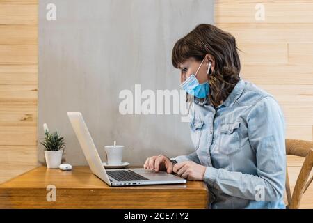 Focused female freelancer in denim shirt and medical mask using earbuds and working remotely in cafe using laptop during coronavirus epidemic Stock Photo