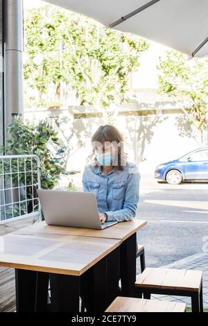 Woman in medical mask working remotely on laptop in outdoor cafe Stock Photo