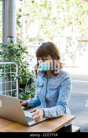 Woman in medical mask working remotely on laptop in outdoor cafe Stock Photo