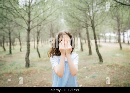 Full body of slim barefoot child in casual dress standing on chair covering face with hands in city park in daylight Stock Photo