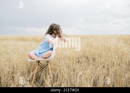 Alone ponder barefoot child with brown hair sitting on chair embracing knees in field with faded grass in overcast weather in autumn and looking away Stock Photo