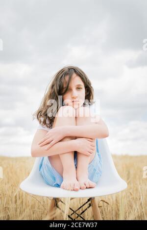 Alone ponder barefoot child with brown hair sitting on chair embracing knees in field with faded grass in overcast weather in autumn and looking at camera Stock Photo