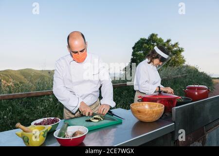 Concentrated man and Woman in medical mask and shield and chef uniforms preparing healthy traditional dishes during workshop in countryside Stock Photo