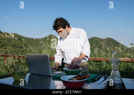 Unrecognizable male chef in uniform and mask checking recipe on laptop while preparing traditional dish lunch in outdoor kitchen during training in countryside Stock Photo