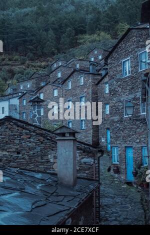 Exterior of aged masonry building with windows and wooden door cobblestone street and cat walking on street with mountains behind under cloudy sky Stock Photo