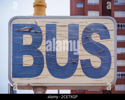 Shabby weathered rectangular road sign indicating bus stop point with word bus written with blue letters on white background and placed against tall modern building in daytime Stock Photo