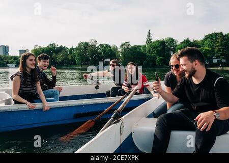 Positive laughing young friends chilling on boats and browsing mobile phone while spending weekend together on peaceful tranquil pond in Saint George, Transylvania, Romania Stock Photo