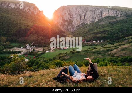 Group of friendly people relaxing on lawn in highlands at sunset and taking selfie on smartphone during vacation in Transylvania Stock Photo