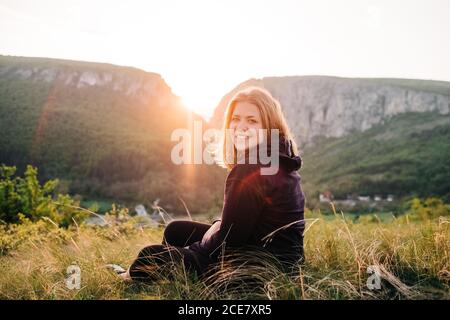 Back view of cheerful female tourist sitting on grass resting in highlands during summer holiday in Transylvania Stock Photo