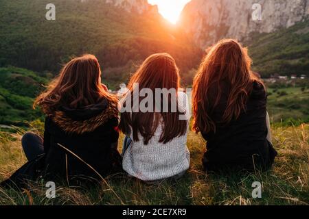 Back view of anonymous female friends chilling on grass in mountains during picturesque sunset in Transylvania and looking over shoulder Stock Photo