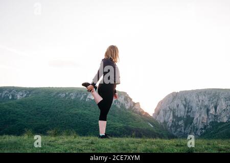 Side view low angle of unrecognizable female in activewear standing on grass and stretching legs while warming up before training and admiring amazing view of highland area Stock Photo