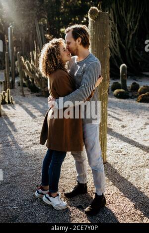 Side view of cheerful man and Woman kissing and embracing each other while spending summer day on blurred background of cactuses in park in Barcelona, Spain Stock Photo