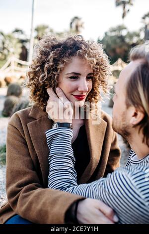 Side view of cheerful man and Woman embracing each other sitting on the floor while spending summer day on blurred background of cactuses in park in Barcelona, Spain Stock Photo