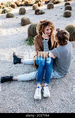 High angle view of cheerful man and Woman smiling and embracing each other sitting on the floor while spending summer day on blurred background of cactuses in park in Barcelona, Spain Stock Photo