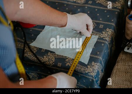 High angle back view of crop anonymous female artisan in latex gloves measuring piece of cloth while manufacturing protective masks during coronavirus pandemic Stock Photo