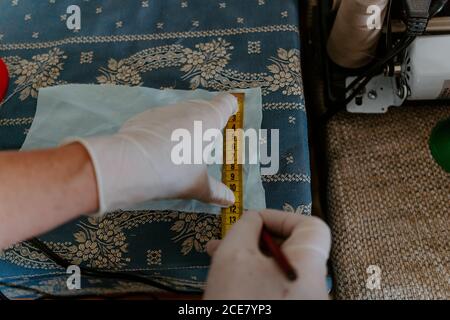 High angle back view of crop anonymous female artisan in latex gloves measuring piece of cloth while manufacturing protective masks during coronavirus pandemic Stock Photo