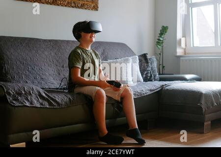 Adorable little boy experiencing virtual reality in modern goggles while playing on sofa with gamepad at home Stock Photo