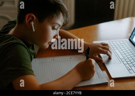 Side view of focused little boy in wireless earphones using laptop and writing down information while doing homework assignment Stock Photo