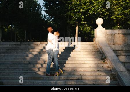 Full length positive diverse couple in casual outfits hugging on staircase on street and looking at each other with love in evening in Madrid Spain Stock Photo