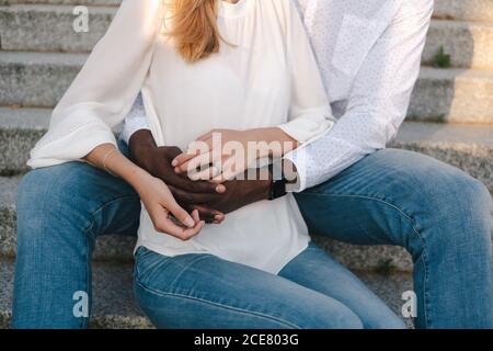 Crop faceless multiethnic couple bonding and holding hands together while African American boyfriend sitting on street stairs behind girlfriend Stock Photo