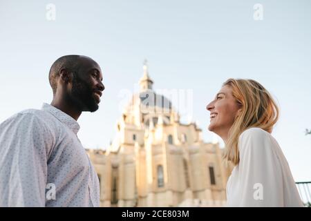 Side view happy smiling multiethnic couple in white shirts standing on street and looking at each other against old architecture in Madrid Stock Photo
