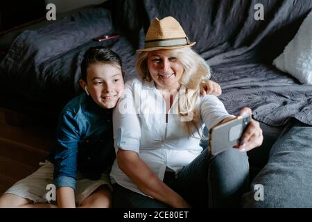 Happy laughing stylish mother and son taking selfie on smartphone while resting on comfy sofa in light living room Stock Photo