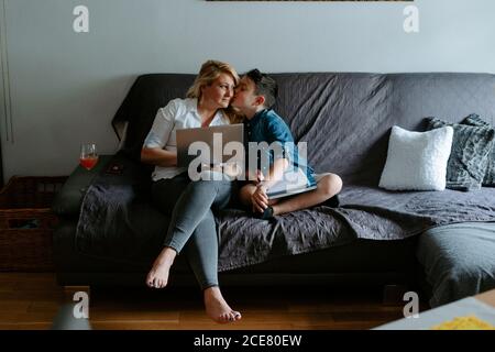 Happy smiling mother with glass of cocktail using netbook while sitting near content son with sheet of paper while spending free time together Stock Photo