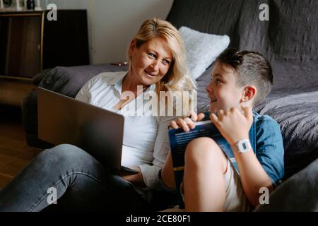 Happy smiling mother with glass of cocktail using netbook while sitting near content son reading book while spending free time together Stock Photo