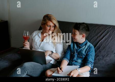 Happy smiling mother with glass of cocktail using netbook while sitting near content son with sheet of paper while spending free time together Stock Photo