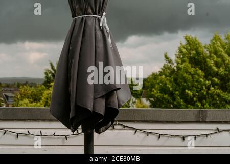 Closed black garden umbrella on terrace against leaden dark cloudy sky before heavy rain on overcast summer day Stock Photo