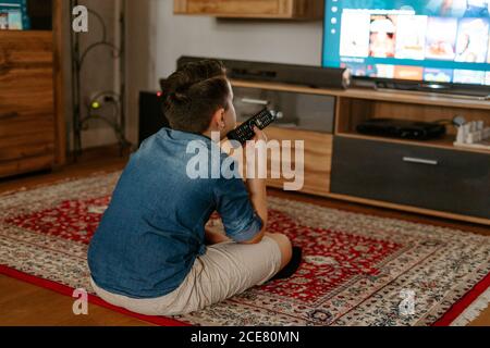 From behind unrecognizable boy in casual clothes sitting on carpet on floor with legs crossed and using remote controller while switching channels on modern tv in cozy living room Stock Photo
