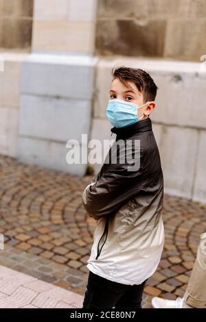 Side view serious boy in warm jacket and medical face mask strolling along paved sidewalk and looking at camera during coronavirus epidemic Stock Photo