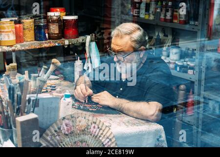 Through glass serious mature male artist in eyeglasses sitting in creative workshop and painting on handmade waver Stock Photo