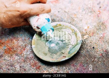 From above crop unrecognizable male artist pouring white paint into weathered palette while working in workshop Stock Photo