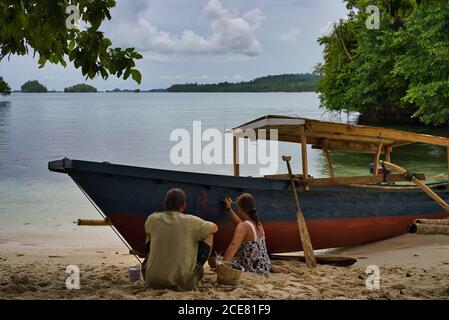 Back view of unrecognizable couple sitting on sandy beach at seaside and writing on boat with red paint Stock Photo