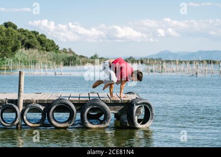 Side view of strong male in casual outfit balancing on arms while performing Kakasana yoga pose during morning training on wooden quay of sea Stock Photo