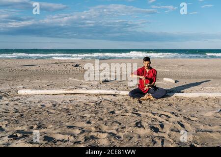 High angle of peaceful young barefooted male in casual clothes sitting on sandy beach in Half Lotus asana near waving ocean and playing Tibetan singing bowl against blue sky Stock Photo