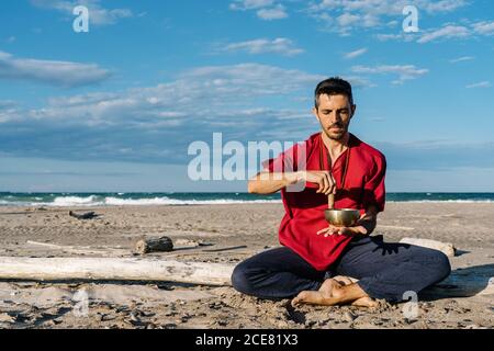 High angle of peaceful young barefooted male in casual clothes sitting on sandy beach in Half Lotus asana near waving ocean and playing Tibetan singing bowl against blue sky Stock Photo