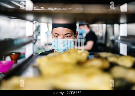 Concentrated female cook in hat and respirator standing against steel pan rack loaded with food while working in modern restaurant kitchen Stock Photo