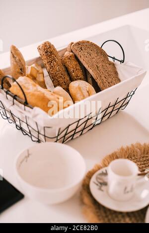 Porcelain tableware set arranged on cloth napkin near basket with fresh bread on table served for breakfast in cozy light apartment Stock Photo