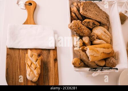 From above of fresh assorted cut bread and buns served in tray and on wooden cutting board on white table during breakfast Stock Photo