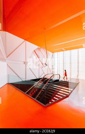 Side view of female walking along spacious passage with stairs and escalator in building in futuristic style with orange floor and ceiling Stock Photo