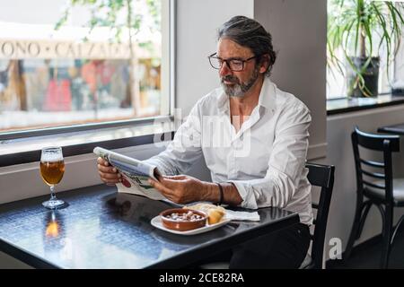 Positive ethnic bearded aged man sitting at table with of glass of foamy beer and reading newspaper near window Stock Photo