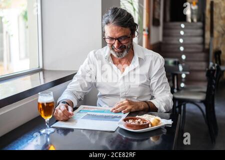 Positive ethnic bearded aged man sitting at table with of glass of foamy beer and reading newspaper near window Stock Photo