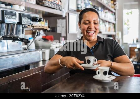 Smiling ethnic female worker in black uniform standing at bar with cups of coffee at daytime Stock Photo
