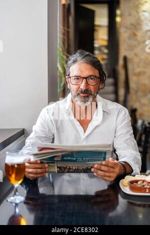 Positive ethnic bearded aged man sitting at table with of glass of foamy beer and reading newspaper near window Stock Photo