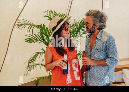 Delighted smiling couple wearing casual summer clothes standing with bottles of beer and bonding in luxury resort tent Stock Photo