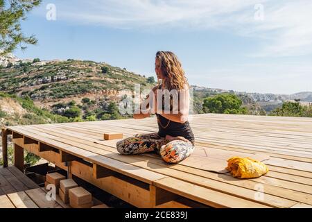 Side view of flexible young female in sportswear sitting on wooden platform in Lotus pose with praying hands while meditating during outdoor yoga practice Stock Photo