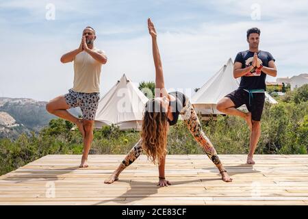Group of full body men and Woman in activewear practicing yoga in Tree pose and wide legged forward bend while standing on mats on wooden platform on nature against tents and plants and mountains Stock Photo