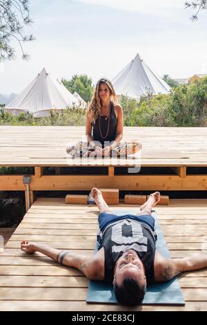 Female yoga trainer sitting in Lotus pose instructing male student relaxing in Corpse asana after finishing outdoor workout on sunny day Stock Photo